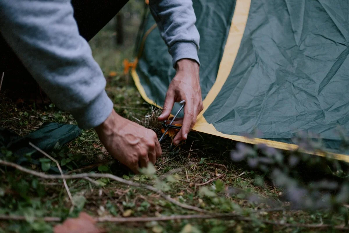 A close-up of a person securing a tent peg in a forested campsite, depicting outdoor camping activities.
