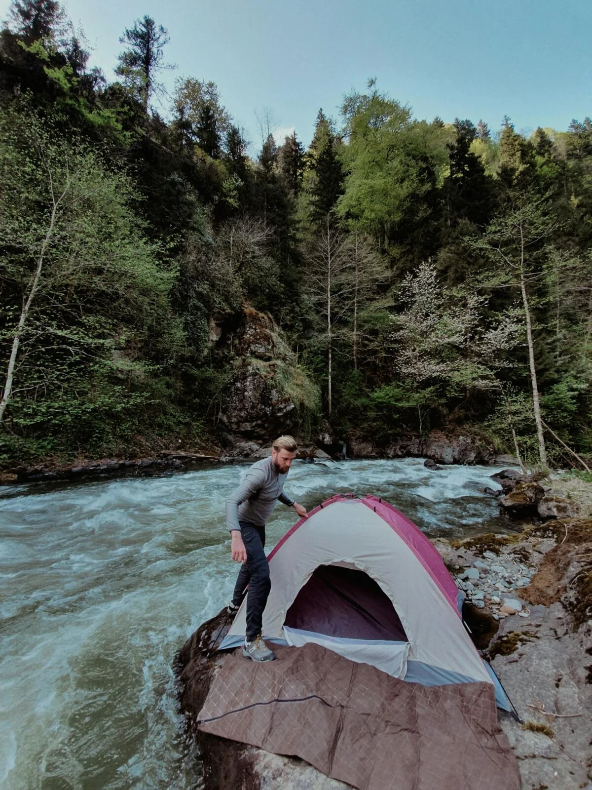 A person setting up a tent beside a rushing river amidst a dense forest.