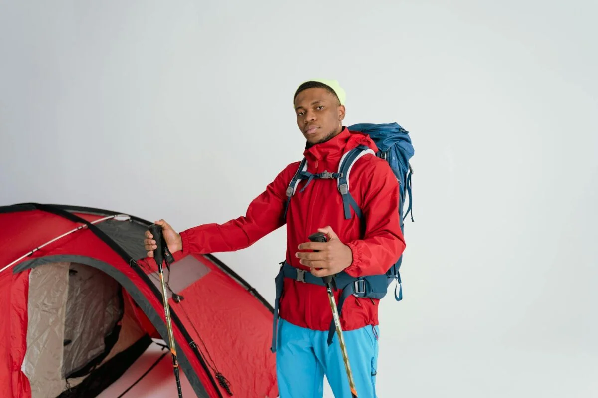 Male hiker in red jacket with backpack and ski poles beside a tent, ready for adventure.